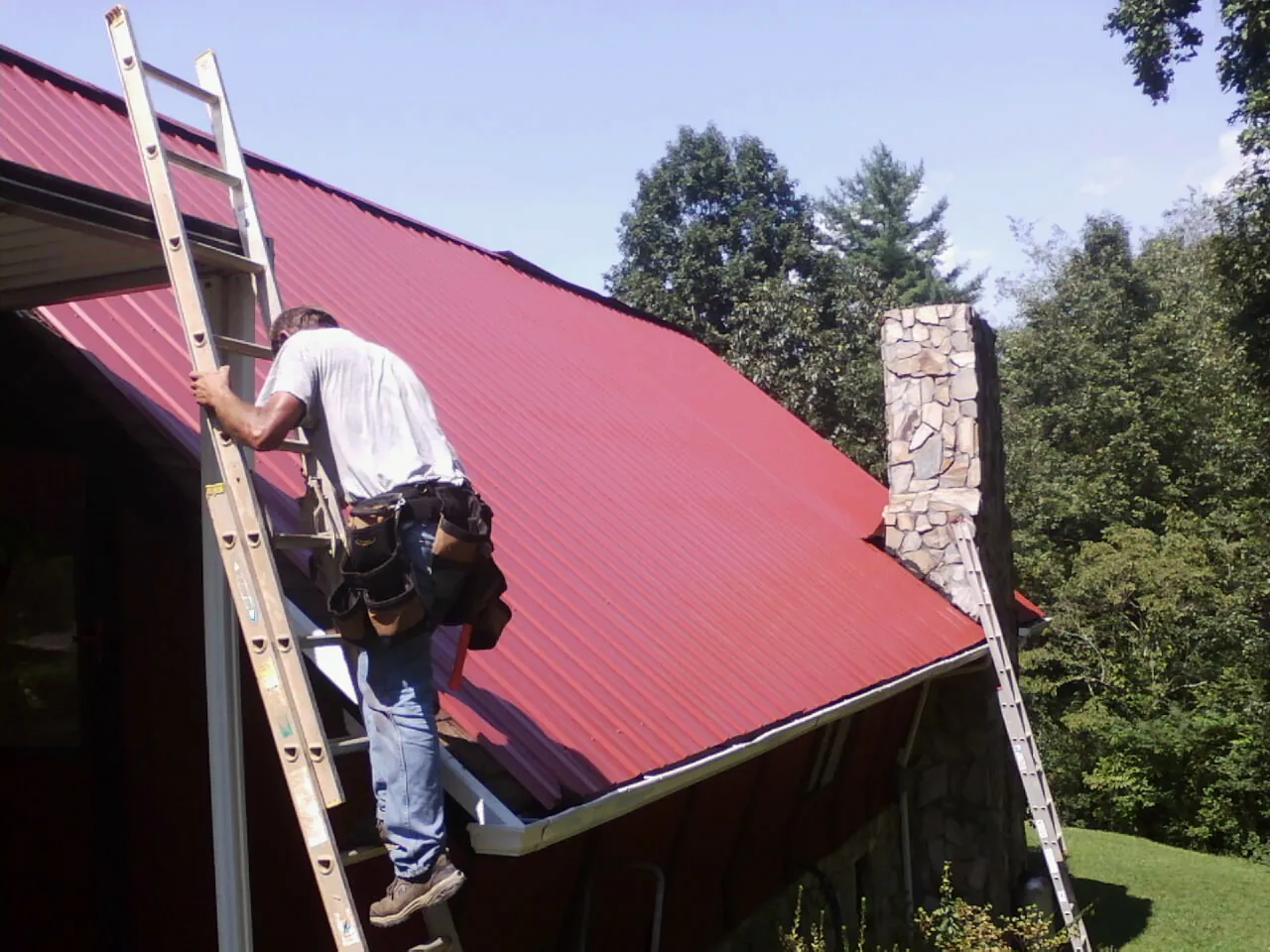 Roofer on a ladder beside a red metal roof and stone chimney — Soffit & Fascia Repair by Roofing Brothers in Talisheek